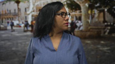Gözlüklü ve mavi bluzlu bir kadın, tarihi binalarla dolu işlek bir caddede başını yana çevirir..