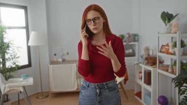 Güzel bir yatak odasında, bebek dekoruyla akıllı telefondan konuşan bir kadın, günlük giysiler içinde kızıl saçlı güzel bir kadın, ev içi ve oyuncaklarla çevrili, ev içi sükuneti taşıyan bir kadın..