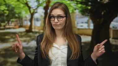 Gözlüklü ve ceketli kadın, ormanda benekli ağaçların arasında kafası karışmış bir ifadeyle omuz silkiyor; şaşkınlık şüphesi belirsizliği.