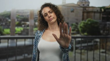 Kadın, bir binanın önündeki balkonda durması için avuç içi kaldırıldığını gösteriyor, beyaz tanktop görünüyor; reddetme sınırı.