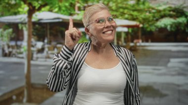 Çene üzerinde eli olan kadın açık kafe yakınlarındaki bir caddede düşünceli poz, kısa saç ve gözlük görünür; merak yansıması.