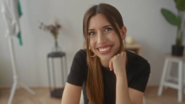 Uzun saçlı, siyah gömlekli İspanyol bir kadın stüdyo odasında çene çalıyor, yanında da bulanık bir bitki var..