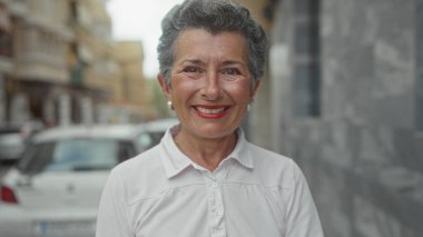Şehir caddesinde gülümseyen gri saçlı yaşlı bir kadın, şehir mimarisi geçmişi olan beyaz bir gömlek giyiyor ve park edilmiş arabalar, açık bir şehir manzarası yaratıyor..