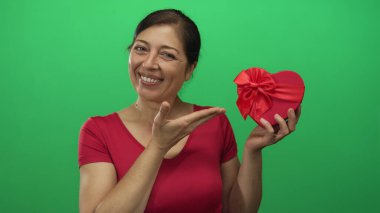 Kadının elinde parlak kırmızı kalp şeklinde bir hediye kutusu ve yeşil stüdyoda parmağıyla kutuya işaret ediyor..