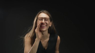 Kahve fincanı tutarken el ele tutuşan ve siyah stüdyo arkaplanında gülümseyen gözlüklü bir kadın..