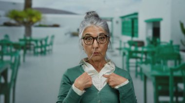 Gözlüklü yaşlı bir kadın, bir restoranın terasında, güneşli bir caddede, yeşil sandalyelerle çevrili bir yerde şaşkınlıkla kendini işaret ediyor..