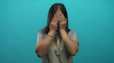 Turkuaz stüdyoda yüzünü kapatan bej önlüklü ve gri tişörtlü genç İspanyol kadın..