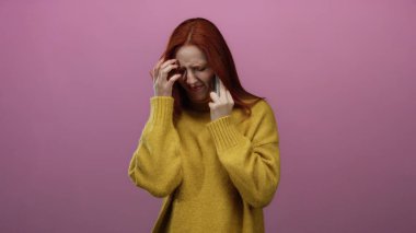 Sarı kazaklı, kırmızı saçlı bir kadın telefonda soyutlanmış pembe arka plana karşı konuşurken hayal kırıklığına uğramış görünüyor. El hareketleriyle ve yüz ifadeleriyle duygularını gösteriyor..