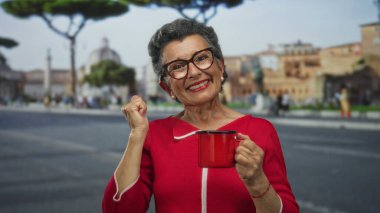 Kırmızı kazaklı son sınıf öğrencisi kadın şehir sokaklarında kahve fincanını tutarken arka planda şehir binaları, açık havada neşe ve neşe ifade ediyor..