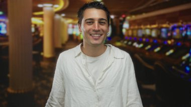 Arka planda kumar makineleri olan bir kumarhanede poz veren genç İspanyol adam, günlük beyaz gömlek giyiyor, canlı kumarhane atmosferini ve şehir gece hayatını ön plana çıkarıyor..