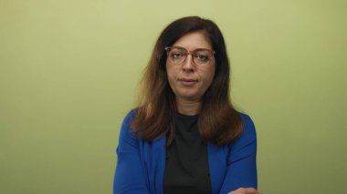 Gözlüklü ve mavi hırkalı bir kadın stüdyoda soluk yeşil duvara karşı kollarını kavuşturuyor; inkar.