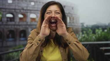 Kahverengi ceketli kadın ellerini ağzının etrafına koyup Roma Kolezyum Binası cephesine bağırıyor..