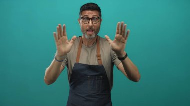 Adam ellerini ve avuçlarını ileri açıp kollarını önlük ve gözlük takmış, deniz mavisi bir stüdyo arkaplanına doğru kaldırırken; sürpriz..