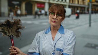 Üst düzey İspanyol kadın üniformalı temizlikçi. Şehir sokağında tozluk tutuyor. Profesyonel hizmeti ve şehir hayatının çabukluğunu yansıtıyor..