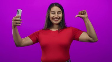 Mor stüdyoda akıllı telefon ve başparmağıyla göğsü gösteren bir kadın; kendini pazarlama güveni.