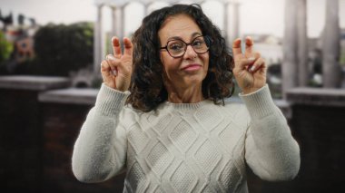 Kıvırcık saçlı, gülümseyen ve el hareketi yapan bir kadın şehir merkezinde neşeli bir atmosfer sergileyen binaların bulanık arka planına sahip..