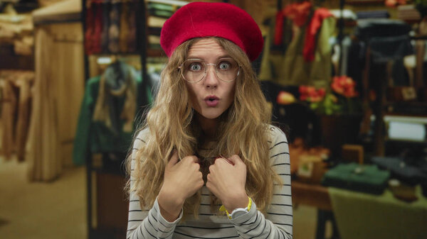 Woman grips hands in surprise inside store as blonde young shopper adjusts glasses under a beret atop striped clothing.