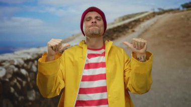 Sarı yağmurluklu ve kırmızı çizgili gömlekli genç adam güneşli bir şehir sokağında kendini gururla işaret ediyor..