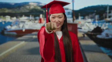 Kırmızı mezuniyet elbisesi giymiş bir kadın, arka planda parlak güneş ışığı altında görünen teknelerle sahil limanında kutluyor, ilham verici ve neşeli bir an yaşatıyor..