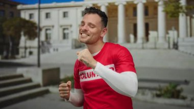 Arka planda tarihi mimarisi olan eski bir üniversite kampüsünün önünde kırmızı gönüllü gömleğiyle neşeyle dans eden genç adam..