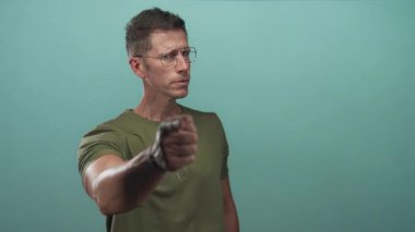 Adam naneli deniz mavisi stüdyosunda, gözlük ve zeytin tişörtü giyen, odaklanmış bakışlar ve sıkılmış çene, çıplak elle izleyiciyi işaret eder..