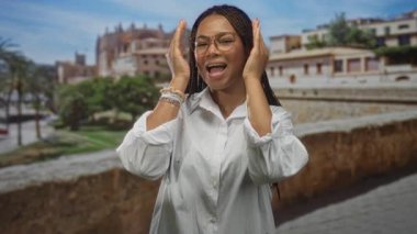 Kadın elleri ile gözlerini kapar ve eski bir kasaba caddesinde taş bir bina tarafından parmaklarının arasından bakar, gözlük ve bileziklerle gülümser; neşe dolu.