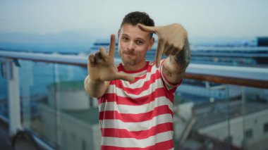 Genç adam çerçeveli bir jest yapıyor, parmakları deniz kenarında bir teknede, çizgili bir gömlek giyiyor, kameraya gülümsüyor, manzaralı bir arka planda gezintiden keyif alıyor..