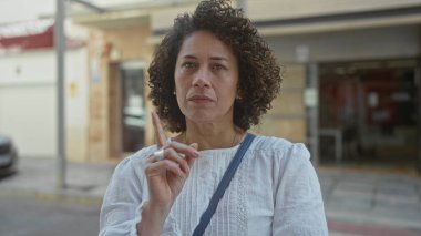 Beyaz bluz giyen ve çapraz-gövde çantası takan kadın, güneş ışığıyla aydınlanan caddede ileriyi işaret ediyor..