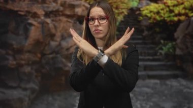 Kırmızı gözlüklü ve siyah ceketli kadın ormanda taş basamaklarda x işareti yaptı; sınır korumasını reddetti.