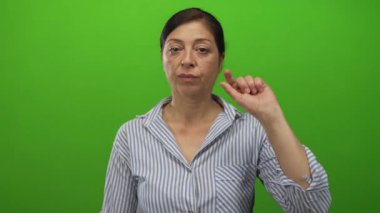 Orta yaşlı İspanyol kadın yeşil stüdyo ortamında parmağı ve işaret parmağıyla ezik el işareti yapıyor..