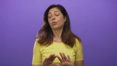 Orta yaşlı İspanyol kadın omuzlarını silkiyor gözleri canlı mor bir stüdyo arka planına karşı kapalı sarı bir bluz giyiyor; kayıtsızlık.