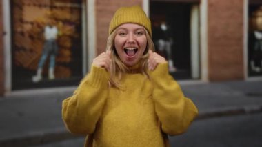 Şehir caddesinde neşeyle gülümseyen bir kadın, rahat sarı bir kazak ve bere giyiyor, açık hava ortamında neşeli bir enerjiyle sonbahar titreşimlerini somutlaştırıyor..