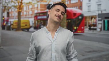 Kısa saçlı genç adam kırmızı otobüsler ve arka planda dükkanlarla dolu işlek bir caddede duruyor. Şehir havası ve rahat bir atmosfer sergiliyor..