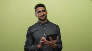 Genç adam yeşil arka plan üzerinde tablet kullanıyor, gözlük takıyor, iki eliyle cihazı tutarken odaklanmış görünüyor, teknoloji ve bağlantı konseptini sunuyor..