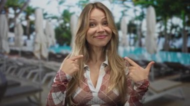 Ekose gömlek giyen gülümseyen sarışın kadın güneşli havuzda şezlongda otururken işaret parmaklarıyla yüzünü gösteriyor..