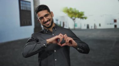 Gülümseyen ve kalp şeklini oluşturan bir adam elleri arka planda bulanık binalar ve ağaçlar olan bir sokakta, sevgi ve sıcaklık saçar..