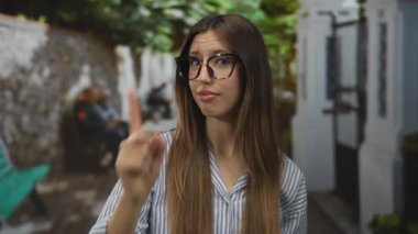 Çizgili mavi gömlekli ve gözlüklü genç İspanyol kadın taştan duvarları ve yeşilliği olan bir şehir caddesini işaret ediyor..