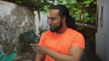 Adam, parlak turuncu bir gömlek ve at kuyruğu giyerken taş bir duvar ve bank yakınında bir caddenin kenarını işaret eder..