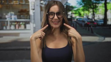 Kadın, gözlük takmış, bir kafenin önündeki caddede gülümsemek için parmaklarını işaret ediyor..
