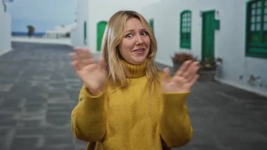 Şehir caddesinde, beyaz binaları ve yeşil kapıları olan sarı süveter giyen bir kadın, açık havada anlamlı bir görünüm sergiliyor..