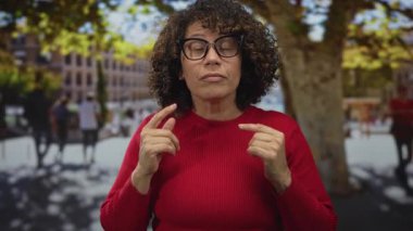 Elleri göğsünde İspanyol bir kadın işlek şehir caddesinde parlak güneş ışığında yapraklı ağaçların altında duruyor..