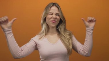 Sarışın kadın, parmaklarını turuncu renkli stüdyo duvarına yaslayarak göğsünü işaret ediyor..