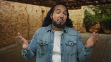 Stüdyoda taş duvar ve ahşap zemin ile hazırlanmış kot ceket ve sakal giyen, elleri göğsüne kenetlenmiş bir adam..