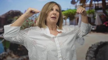 Beyaz bluzlu orta yaşlı kadın, binadaki omuz hizasında görünen elleri ile kollarını kaldırıyor; sükunet barış yansıması.