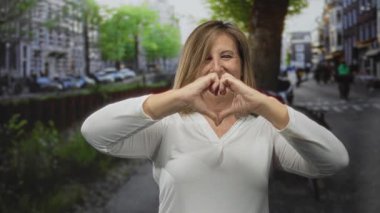 Orta yaşlı beyaz kadın gülümseyen ve kalp oluşturan şehir sokaklarında elleri ile; aşk mutluluk neşeyi etkiler.
