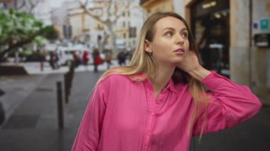 Pembe tişörtlü kadın canlı bir şehir caddesinde arka planı bulanık bir şekilde dikkatlice dinliyor, şehir merkezindeki şehir enerjisini ve merakını yakalıyor..