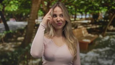 Parkta pembe bluz giyen, işaret parmağı gösteren ve kameraya göz kırpan sarışın bir kadın..