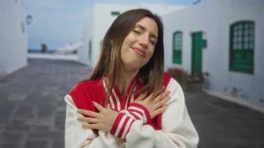 Kırmızı takım ceketli genç bir kadın, güneşli bir sokakta kollarını göğsünün üzerinde kavuşturuyor, gözleri kapalı, kendi rahatı için sükunet sağlıyor..