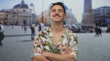 Çapraz kollu ve tropikal tişörtlü genç İspanyol adam, St. Peter Meydanı, Vatikan 'da kendine güvenle gülümsüyor, ikonik bir açık hava ortamında canlı bir turist havası yaratıyor..