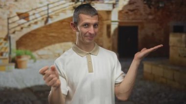Sıradan gömlekli bir adam taşra arka planına karşı jest yapar. Onay ya da sunum ima eder. Elleri işaret eder ve 